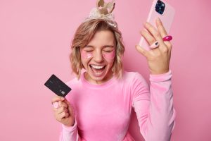 Overjoyed Positive Young Woman Wears Crown Dress Foolishes Around Holds Plastic Card Smartphone Exclaims Loudly Applies Hydrogel Beauty Patchesunder Eyes Isolated Pink Backgroud Scaled